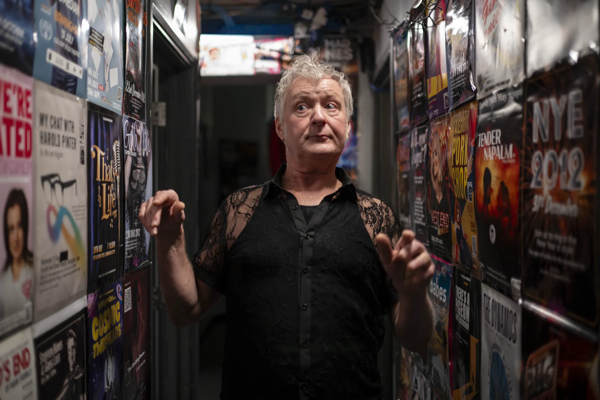 A man in a black lace shirt mid-gesture in the poster-lined corridor